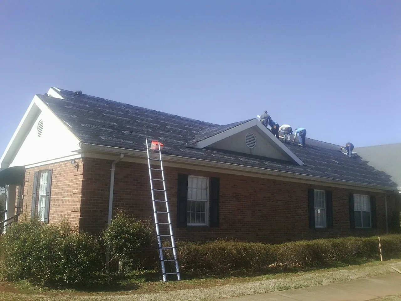 Crew tearing shingles off a brick ranch roof with a ladder set — Metal Roof Repair work by our crew in Wingate